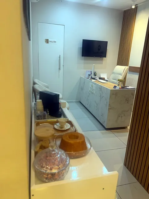 Recepção da clínica, com mesa de atendimento, TV e a porta do consultório